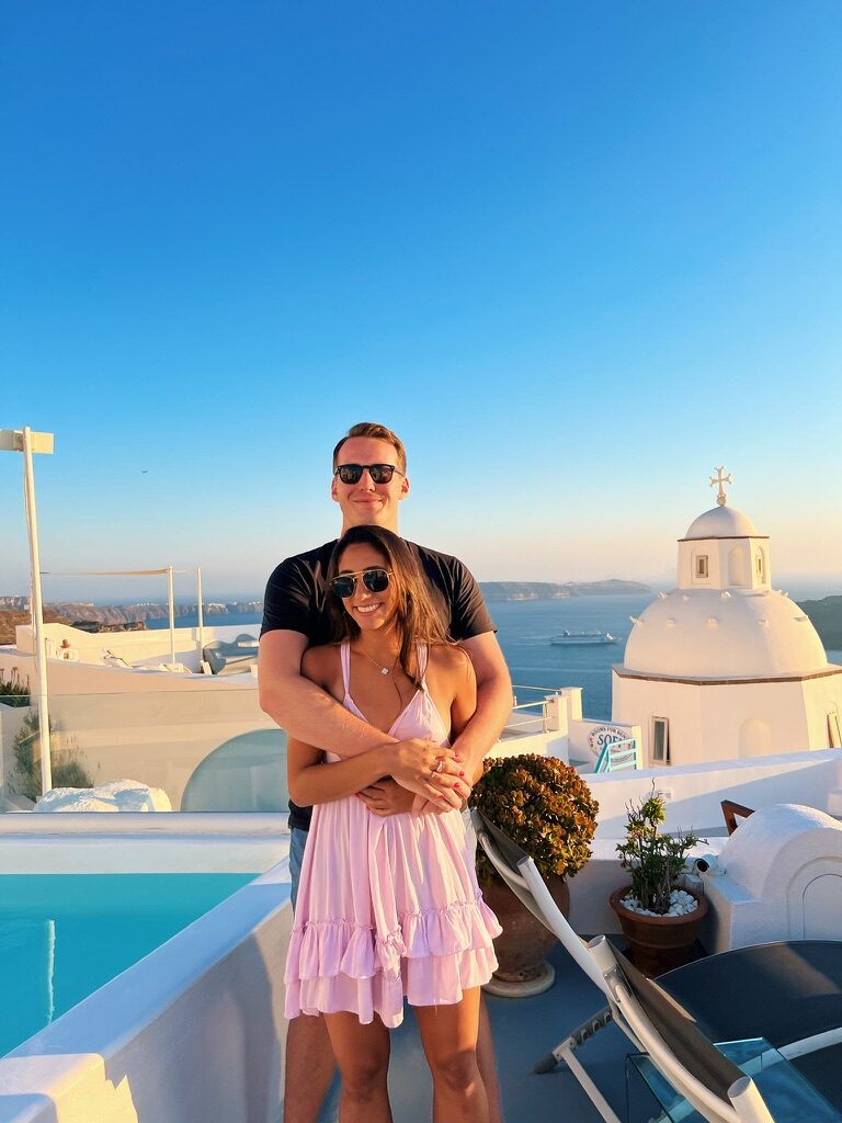 The couple on a terrace at sunset in Santorini, a white domed chapel behind them