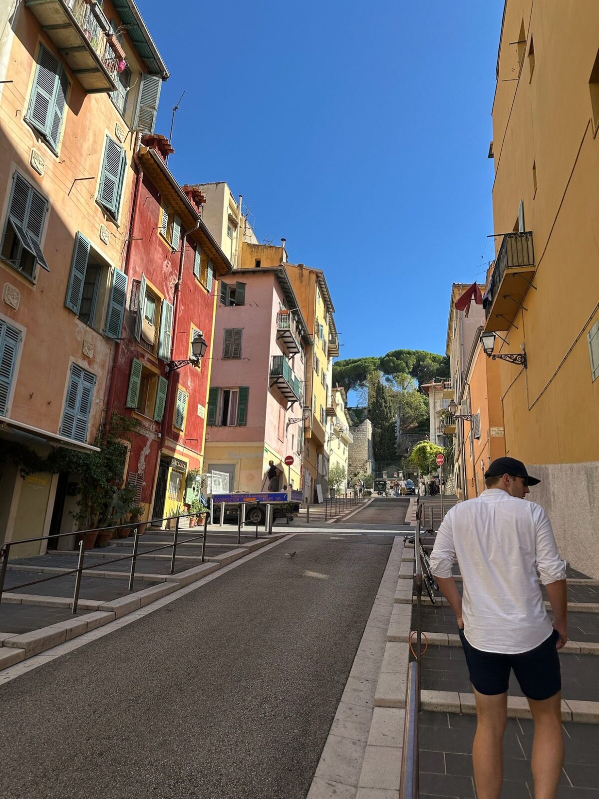 Ryan walking up a street of orange, red and pink buildings with green shutters