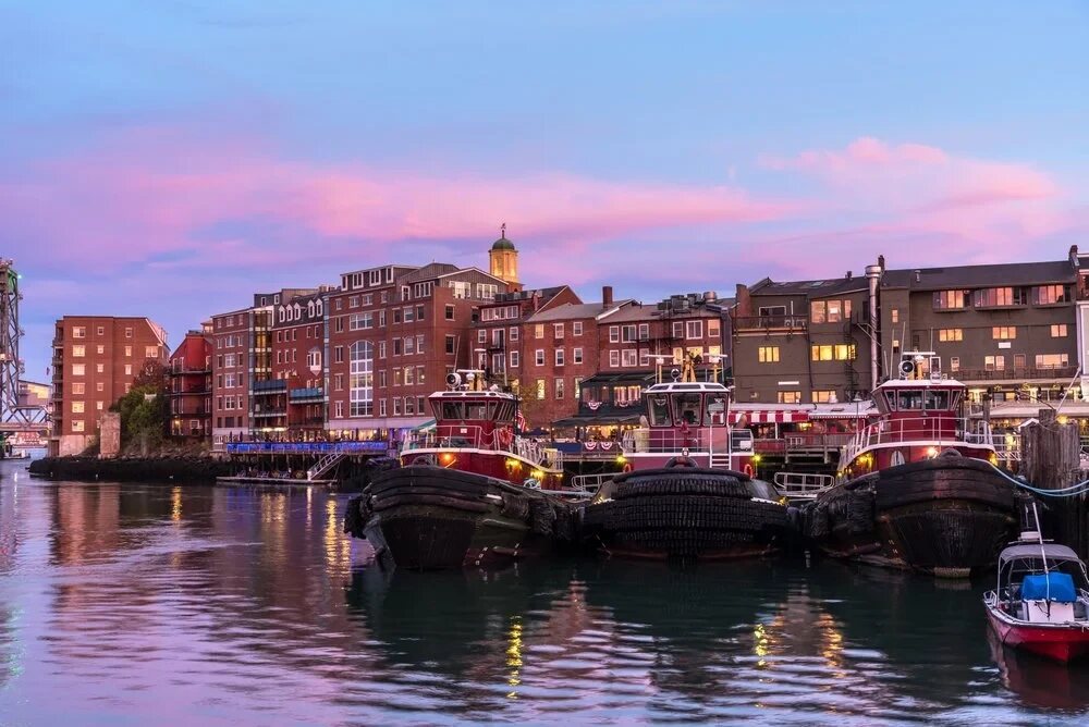Three red tugboats docked along the Portsmouth waterfront under a pink and purple dusk sky