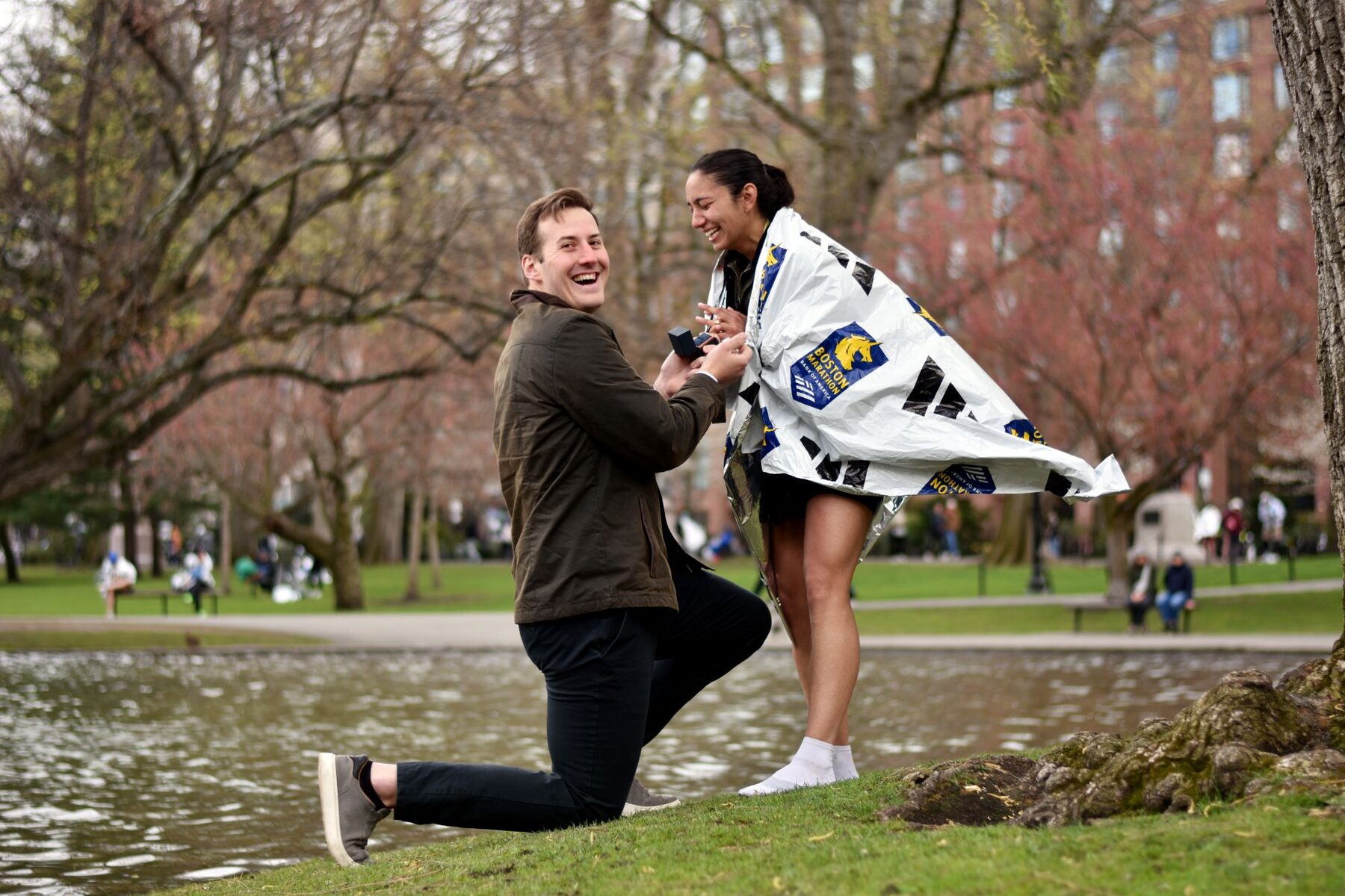 Ryan on one knee beside the pond, laughing, as Maddie, wrapped in a Boston Marathon finisher's blanket, looks at the ring